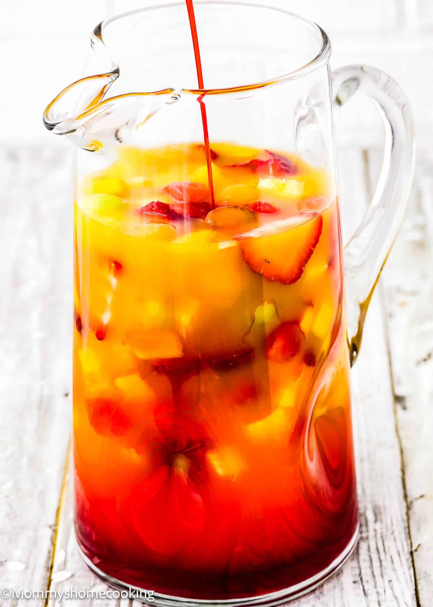 A glass pitcher filled with Venezuelan Tizana (Easy Fruit Punch Mocktail), a vibrant yellow and red drink featuring sliced strawberries and other fruit, with red liquid being poured in.