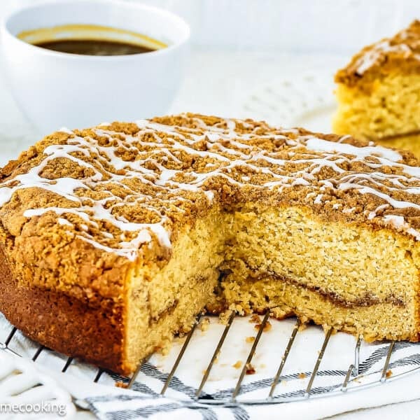 Round Easy Eggless Coffee Cake with crumb topping and white icing drizzle, partially sliced, displayed on a cooling rack with coffee cups and a cake slice in the background.