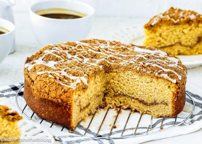 Round Easy Eggless Coffee Cake with crumb topping and white icing drizzle, partially sliced, displayed on a cooling rack with coffee cups and a cake slice in the background.