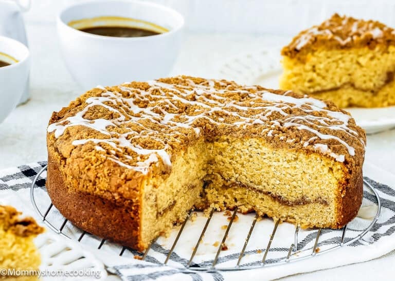 Round Easy Eggless Coffee Cake with crumb topping and white icing drizzle, partially sliced, displayed on a cooling rack with coffee cups and a cake slice in the background.