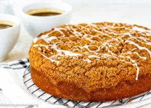 A round Easy Eggless Coffee Cake with white icing drizzle sits on a cooling rack, with two cups of coffee in the background.