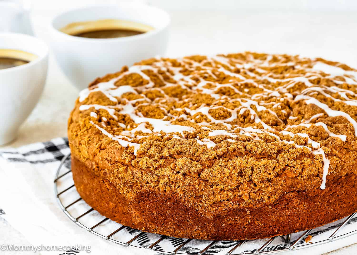 A round Easy Eggless Coffee Cake with white icing drizzle sits on a cooling rack, with two cups of coffee in the background.