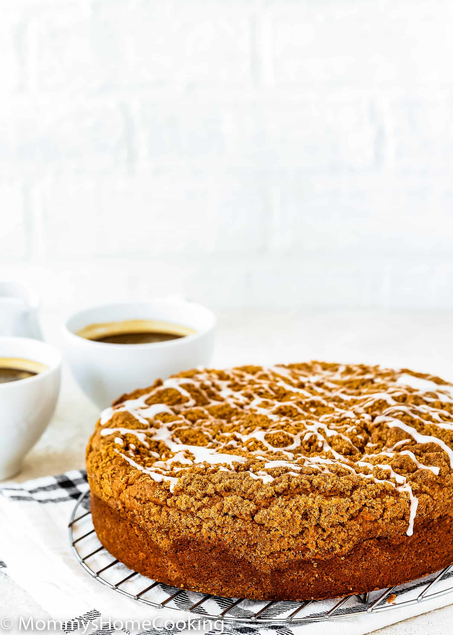 An Eggless Coffee Cake with streusel and glaze on top over a cooling rack with two cup of coffee on the background.