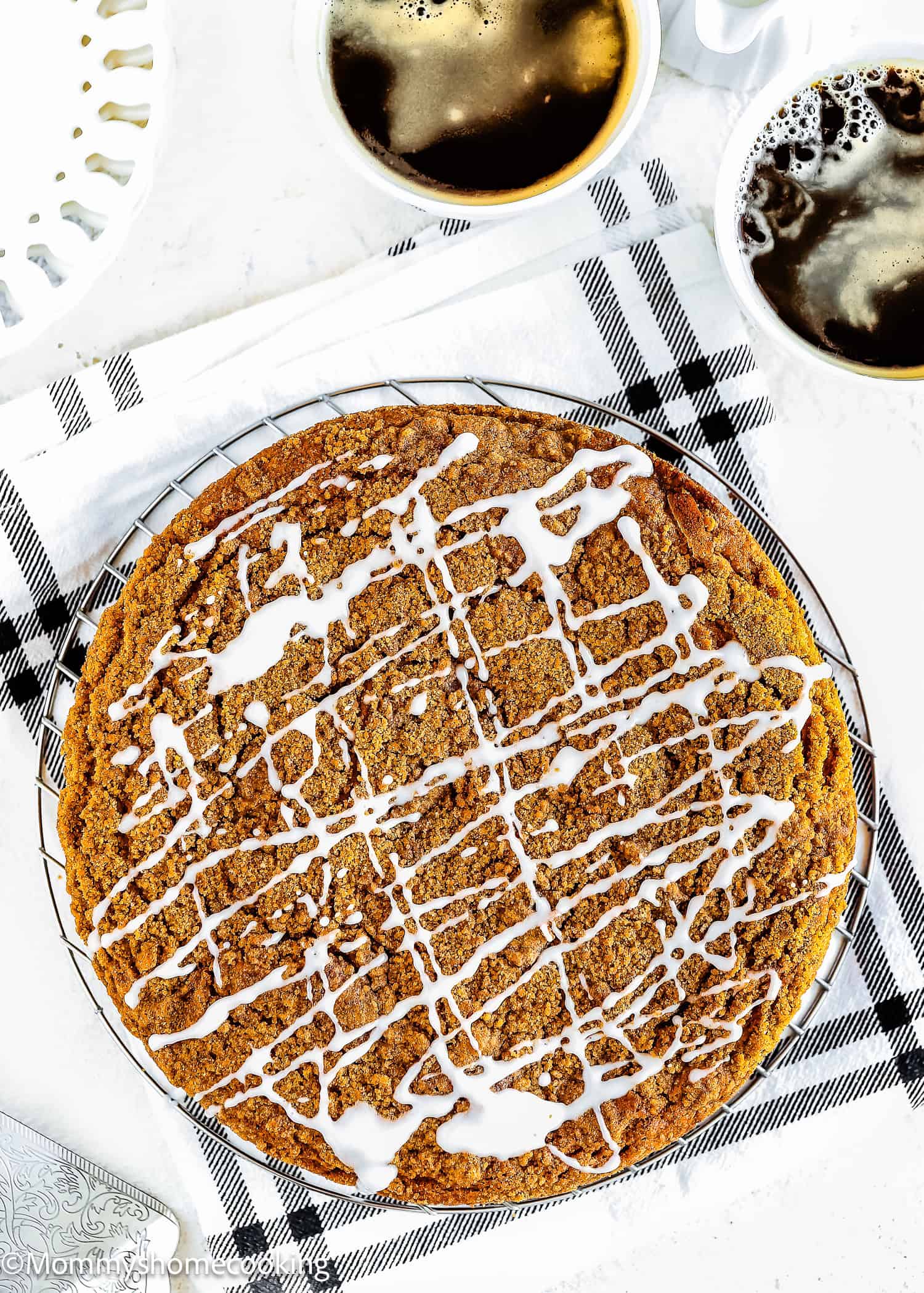 An Easy Eggless Coffee Cake with white icing drizzle sits on a wire rack over a striped cloth, accompanied by two cups of black coffee nearby.