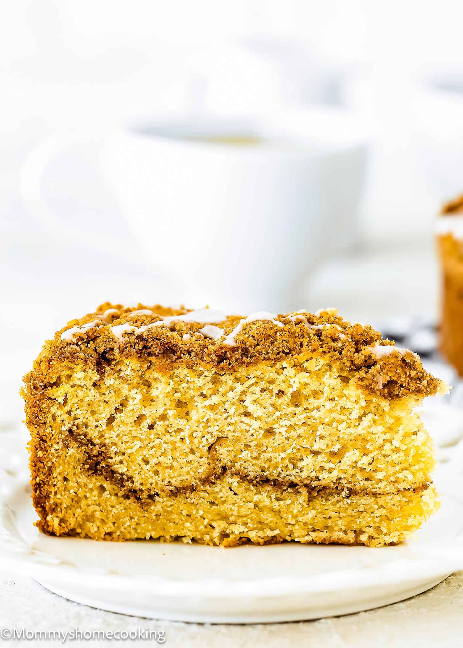 A slice of Easy Eggless Coffee Cake with crumb topping and icing, served on a white plate with a blurred cup in the background.
