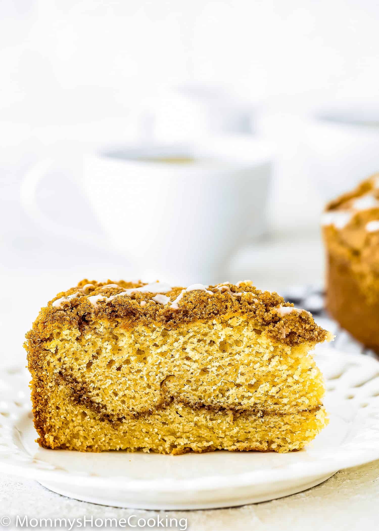 A slice of Eggless Coffee Cake on a plate with a cup of coffee in the background.