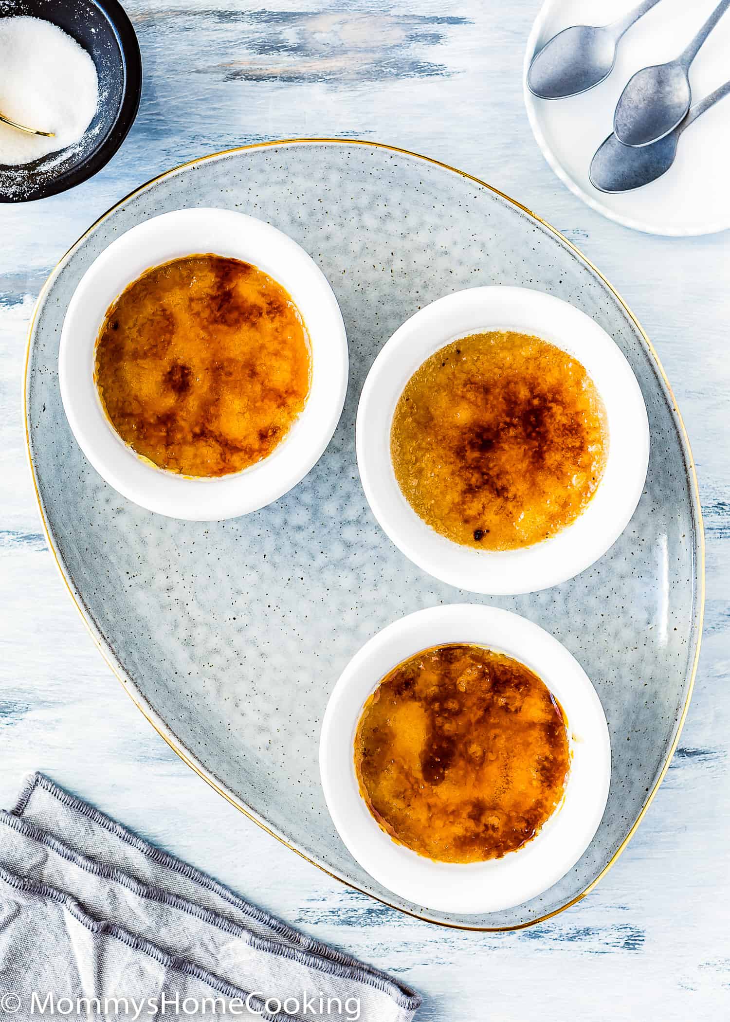 three egg-free creme brulee with a sugar caramelized top over a blue serving plate with a grey kitchen towel on the side and three spoons.