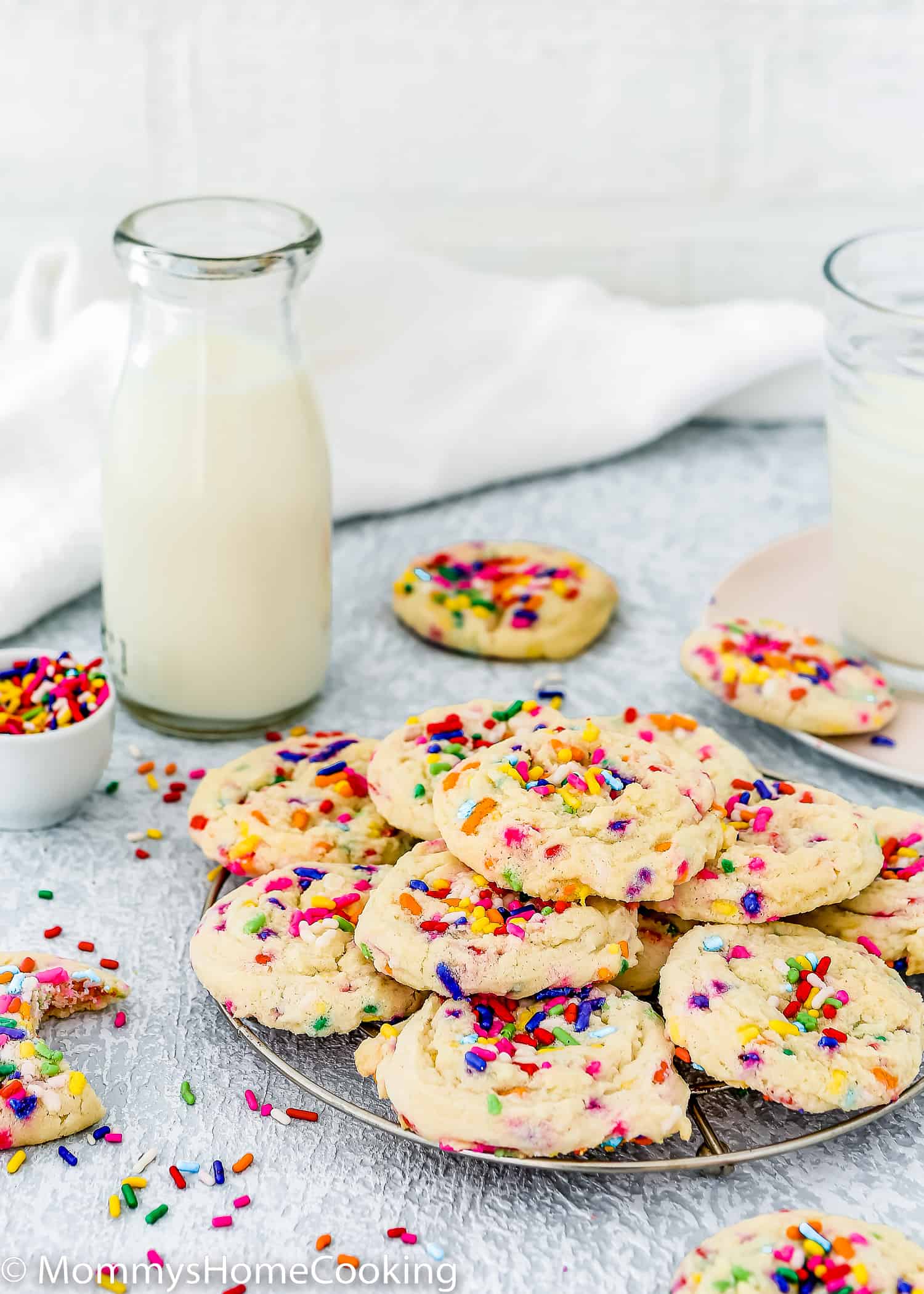 eggless Funfetti cookies on a cooling rack with sprinkles and more cookies and a glass of milk around it.