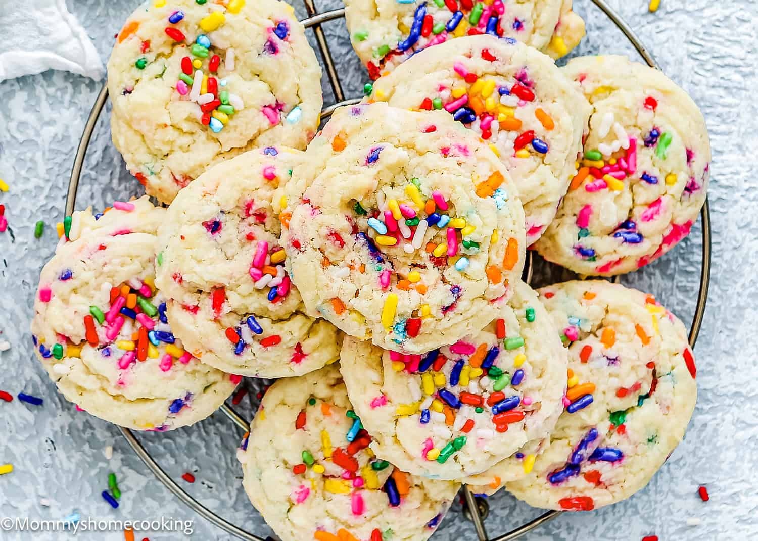 A pile of colorful Eggless Funfetti Cookies displayed on a wire cooling rack. Each cookie is topped with a variety of rainbow sprinkles.