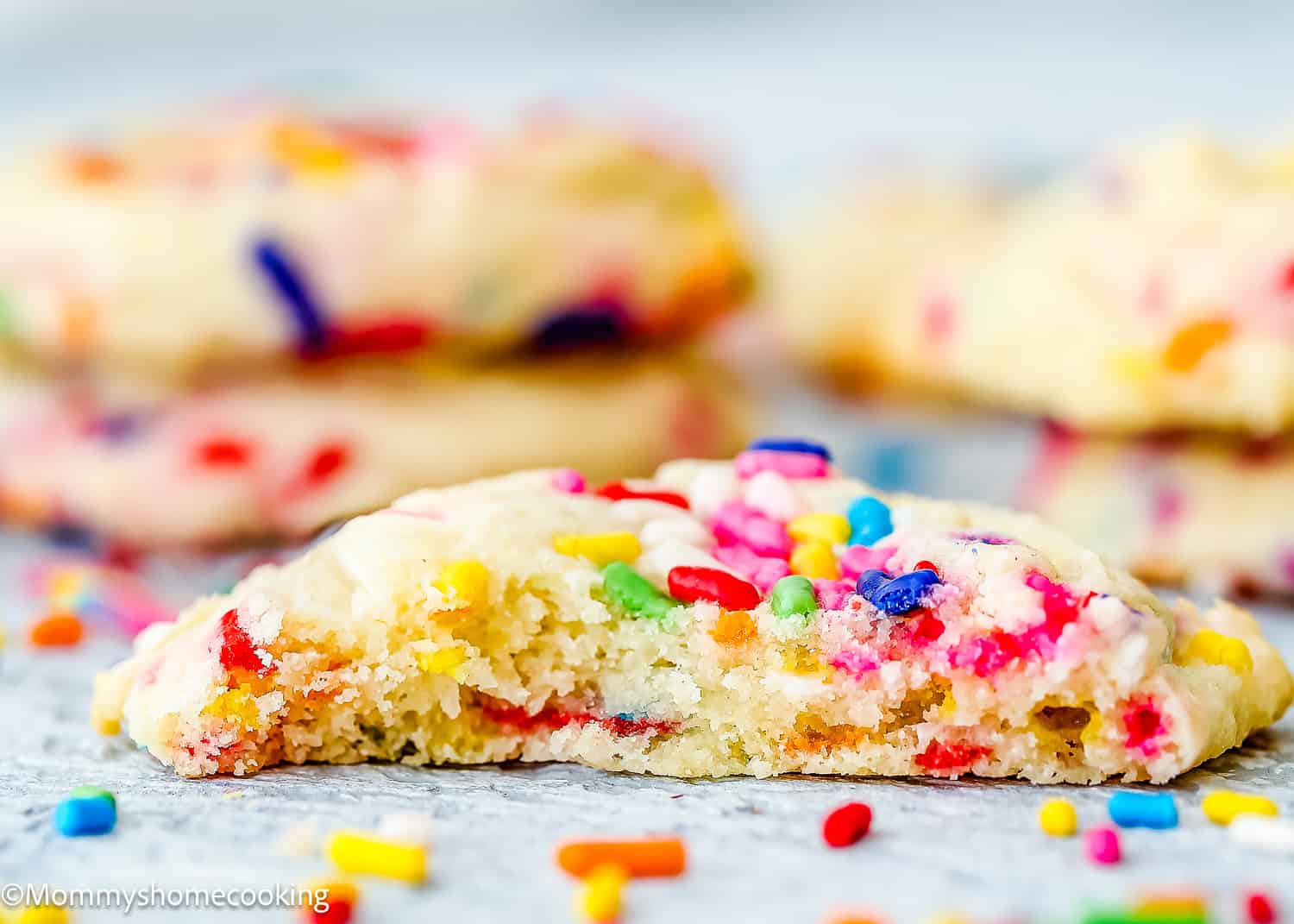 A close-up of a halved Eggless Funfetti Cookie with colorful sprinkles on a light surface, with more cookies and sprinkles blurred in the background.