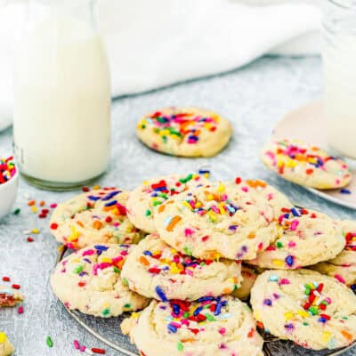 A cooling rack holds colorful Eggless Funfetti Cookies. A glass bottle of milk, a glass of milk, and a plate with a few cookies are placed nearby on a light-colored surface.