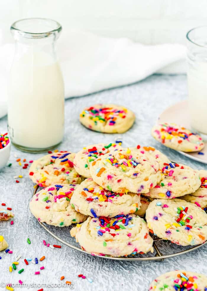 A cooling rack holds colorful Eggless Funfetti Cookies. A glass bottle of milk, a glass of milk, and a plate with a few cookies are placed nearby on a light-colored surface.