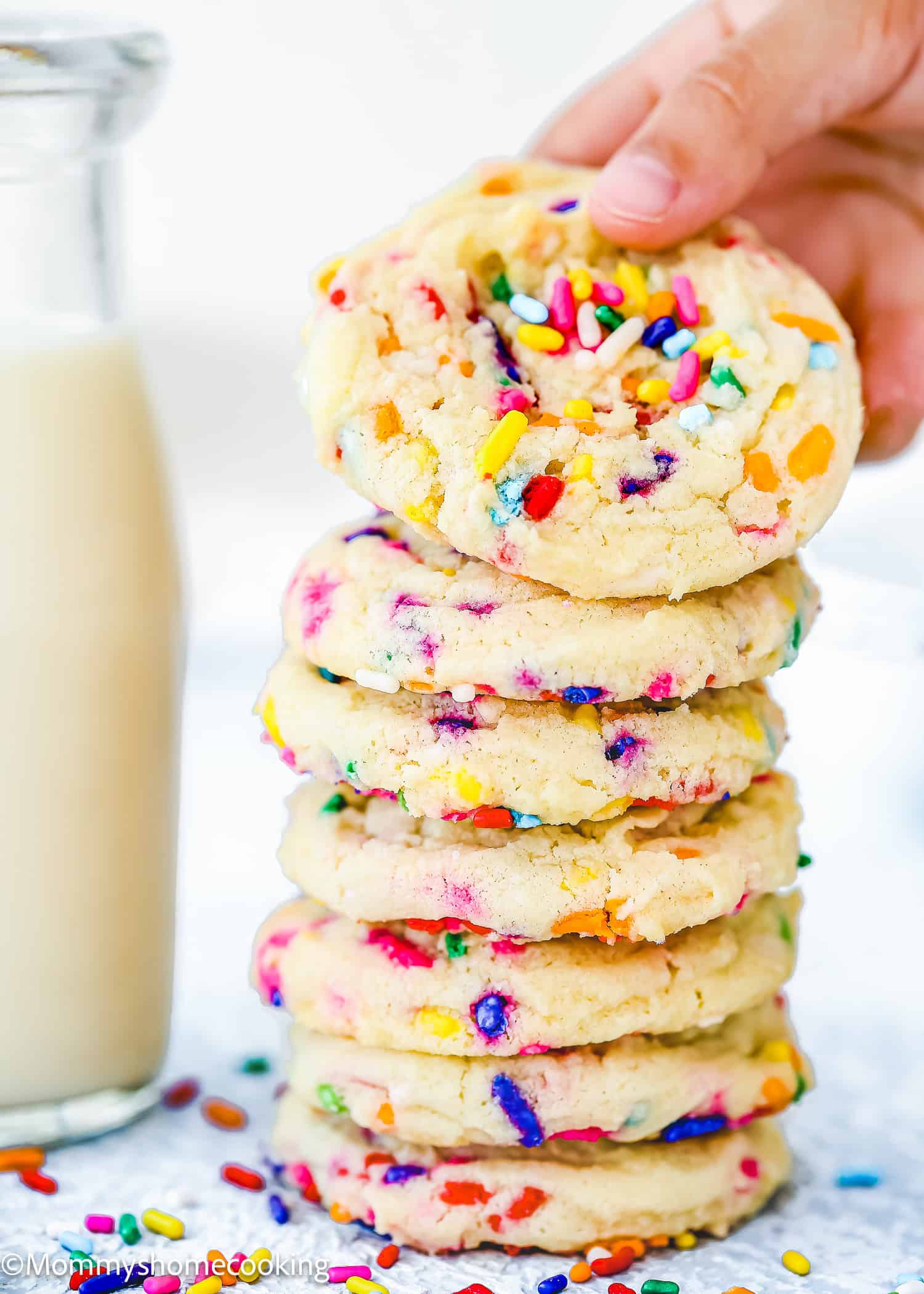 A hand lifts a sprinkle-covered Eggless Funfetti cookie from a stack of similar cookies next to a glass bottle of milk. The cookies display a mix of colorful sprinkles.