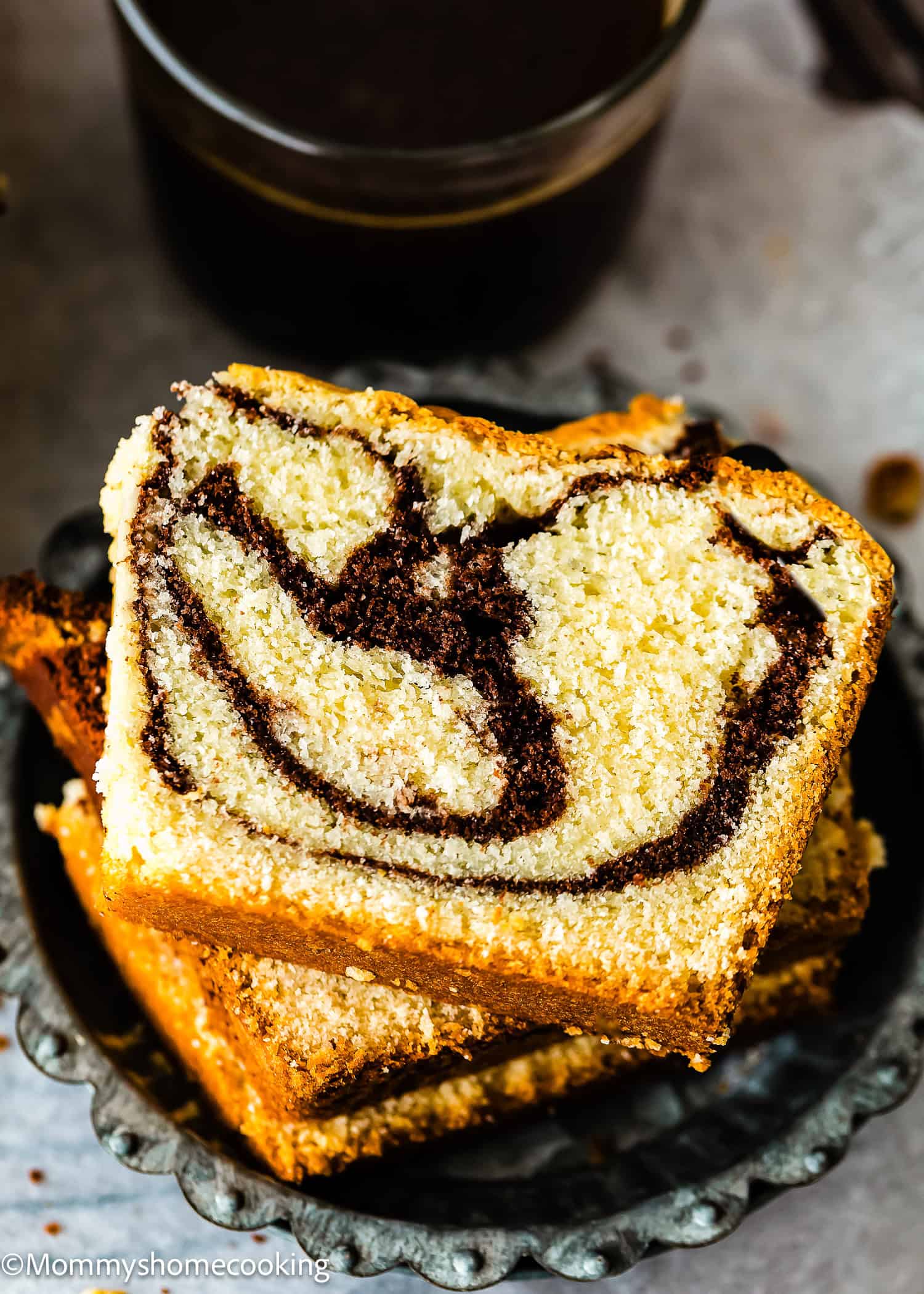 Two slices of eggless marble cake with chocolate and vanilla swirls are stacked on a metal plate, with a dark beverage in the background.