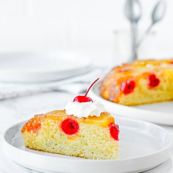 a slice of Eggless Pineapple Upside Down Cake on a plate with whipped cream and the whole cake in the background.