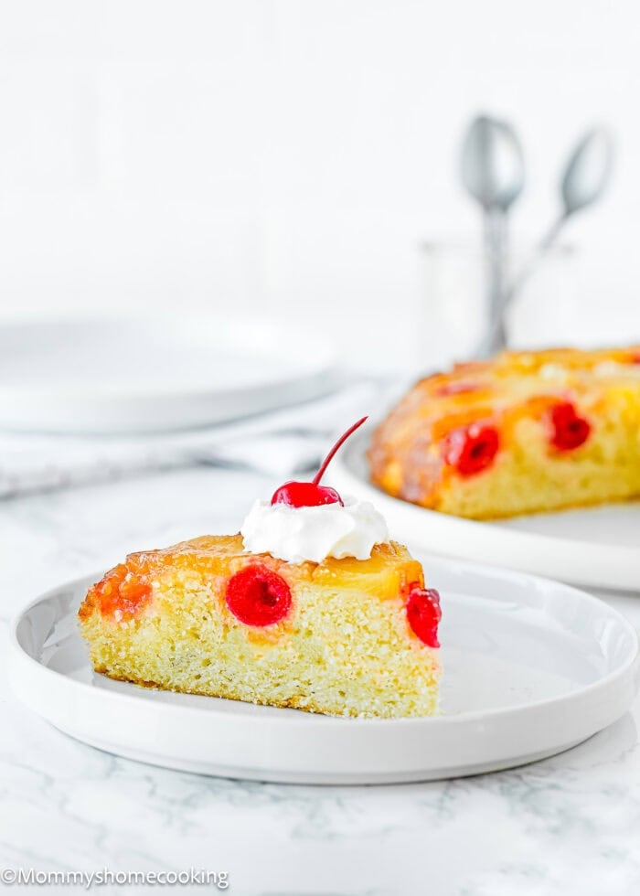 a slice of Eggless Pineapple Upside Down Cake on a plate with whipped cream and the whole cake in the background.