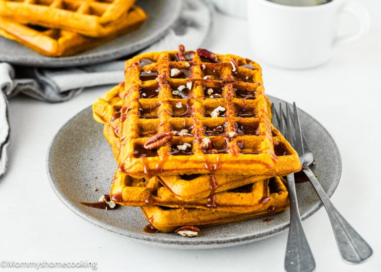 A stack of Quick & Easy Eggless Pumpkin Waffles topped with pecans, syrup, and coarse salt sits on a gray plate with two forks; a coffee cup and extra waffles are in the background.