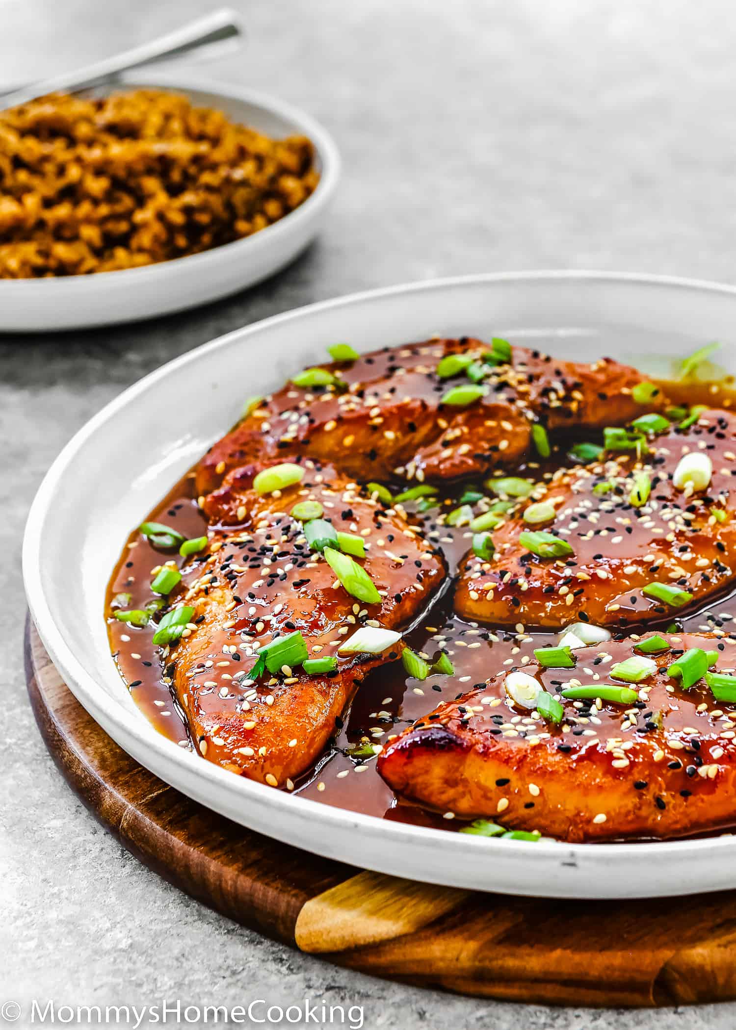 Asian-Style sticky Chicken Breasts in a white skillet garnished with green onion and sesame seeds and a plate of rice in the background.