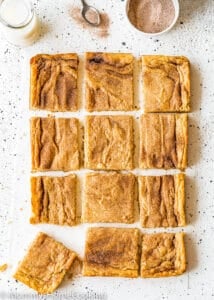 Twelve egg-free Snickerdoodle Bars over a white surface with a bottle of milk on the side and a bowl with cinnamon sugar.