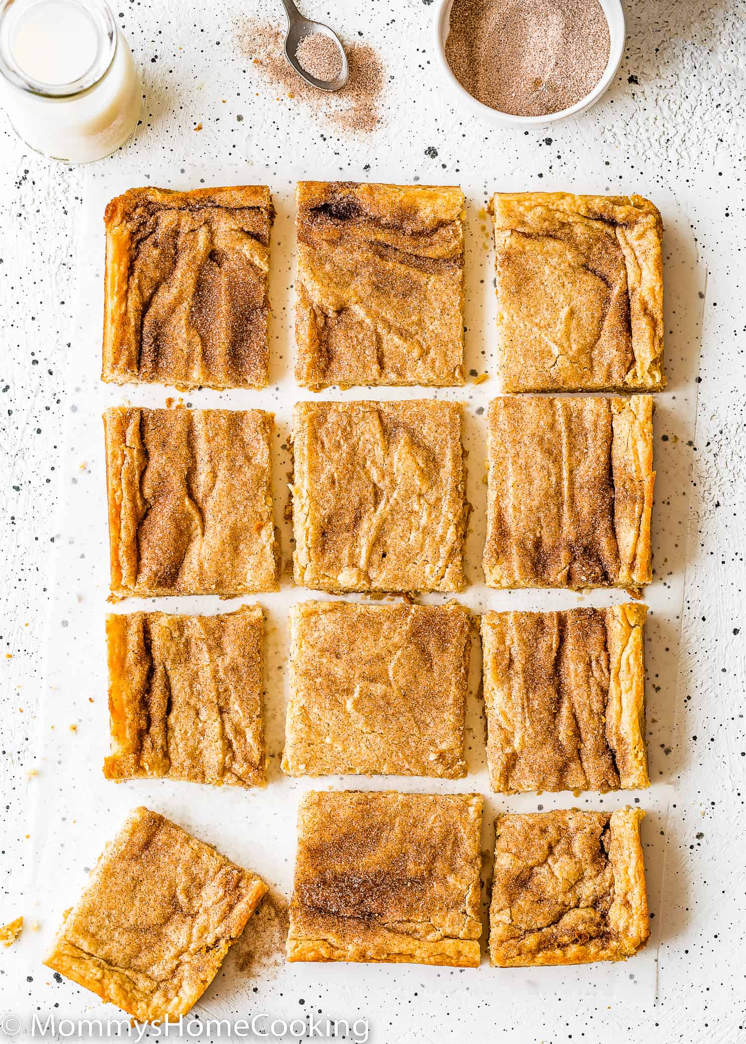 Twelve egg-free Snickerdoodle Bars over a white surface with a bottle of milk on the side and a bowl with cinnamon sugar.