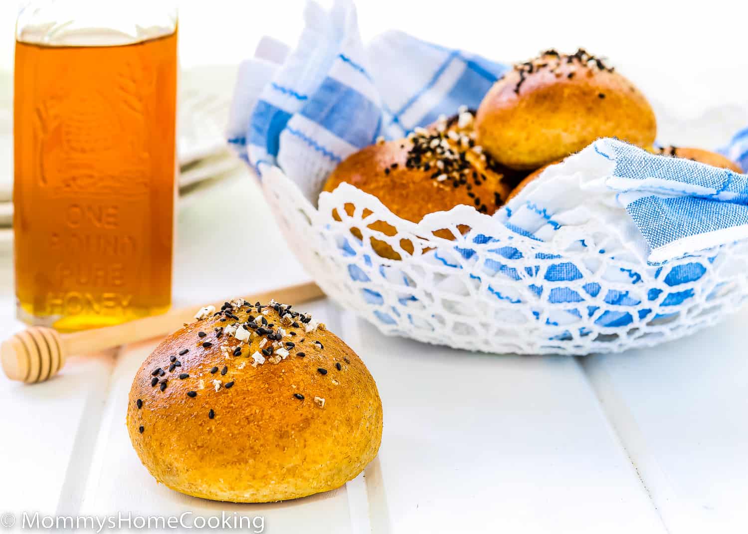 An Eggless Honey Whole Wheat Roll with sesame seeds, pretzel salt, a honey bottle, and a bread basket in the background.
