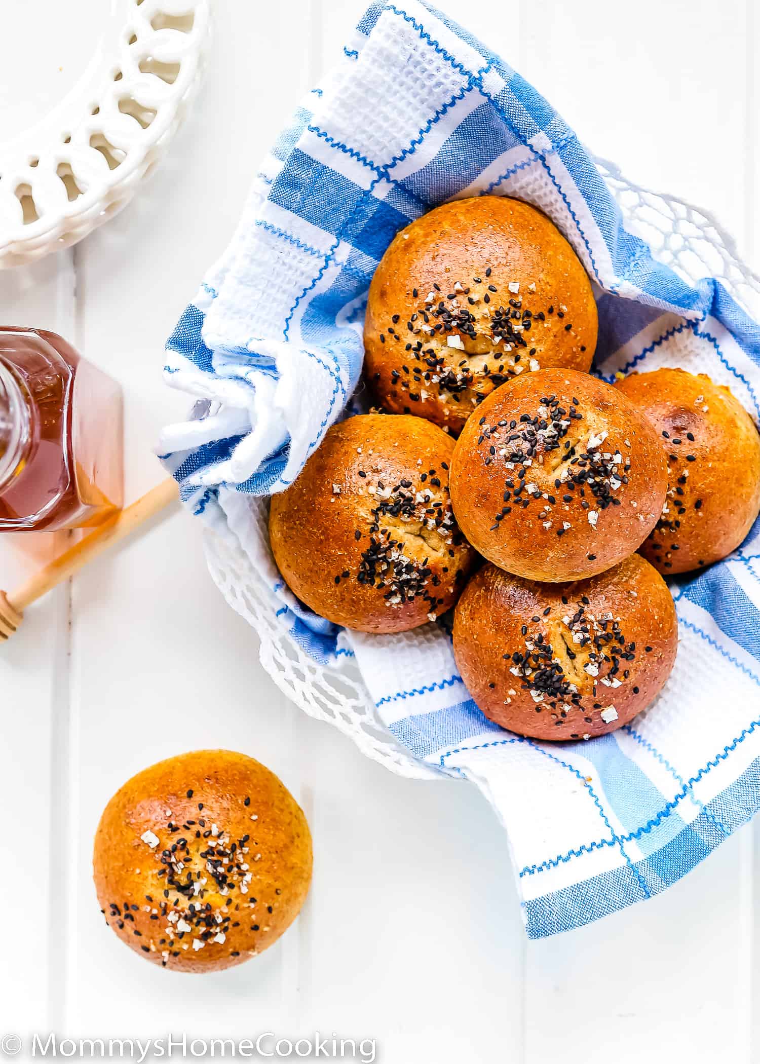 Eggless Honey Whole Wheat Rolls with sesame seeds and pretzel salt in a bread basket with a blue kitchen towel and a bottle of honey on the side.