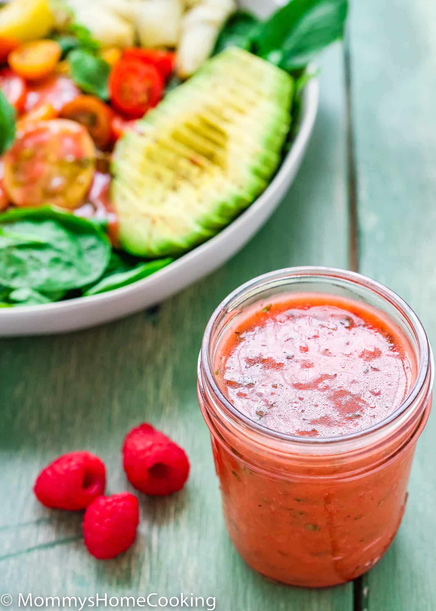 Raspberry Vinaigrette in a glass jar with fresh raspberries on the side and a salad in the background over a green surface.