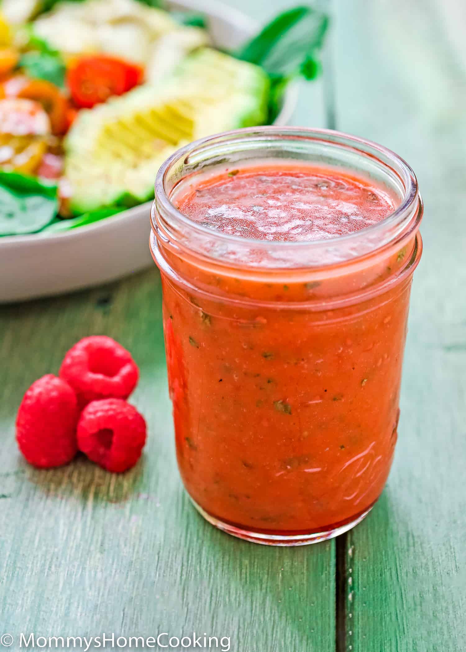 Raspberry vinaigrette in a glass with fresh raspberries on the side and a salad in the background.