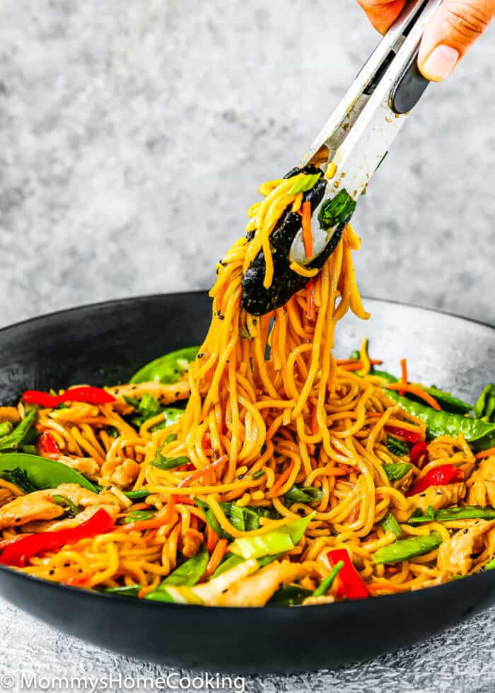 A hand using tongs to pick up Easy Chicken Lo Mein, stir-fried with vegetables in a black bowl.