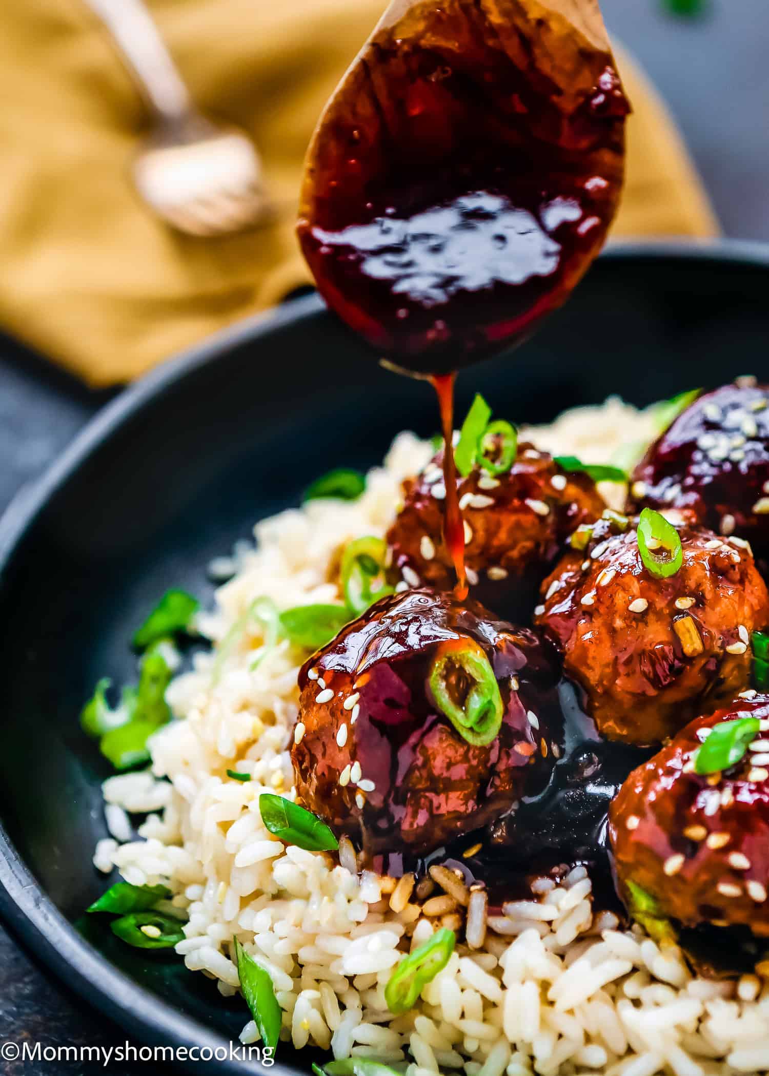 A spoon drizzles sauce over easy Teriyaki turkey meatballs, nestled on rice and garnished with sesame seeds and chopped green onions, all served in a sleek black bowl.