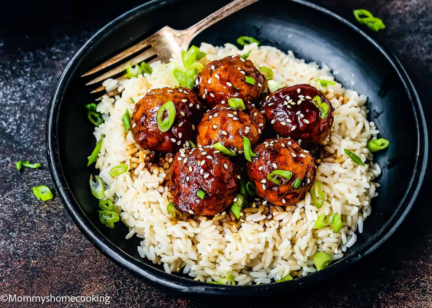 A black bowl filled with steaming white rice is topped with six easy teriyaki turkey meatballs, delectably glazed and egg-free, garnished with chopped green onions and sesame seeds. A fork rests invitingly on the side.