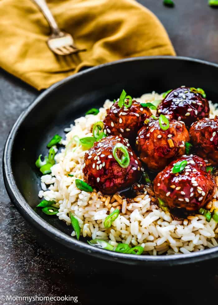 A black bowl showcases Easy Teriyaki Turkey Meatballs, egg-free and glazed to perfection, topped with sesame seeds and green onions on a bed of rice. A fork rests on a folded yellow napkin in the background.