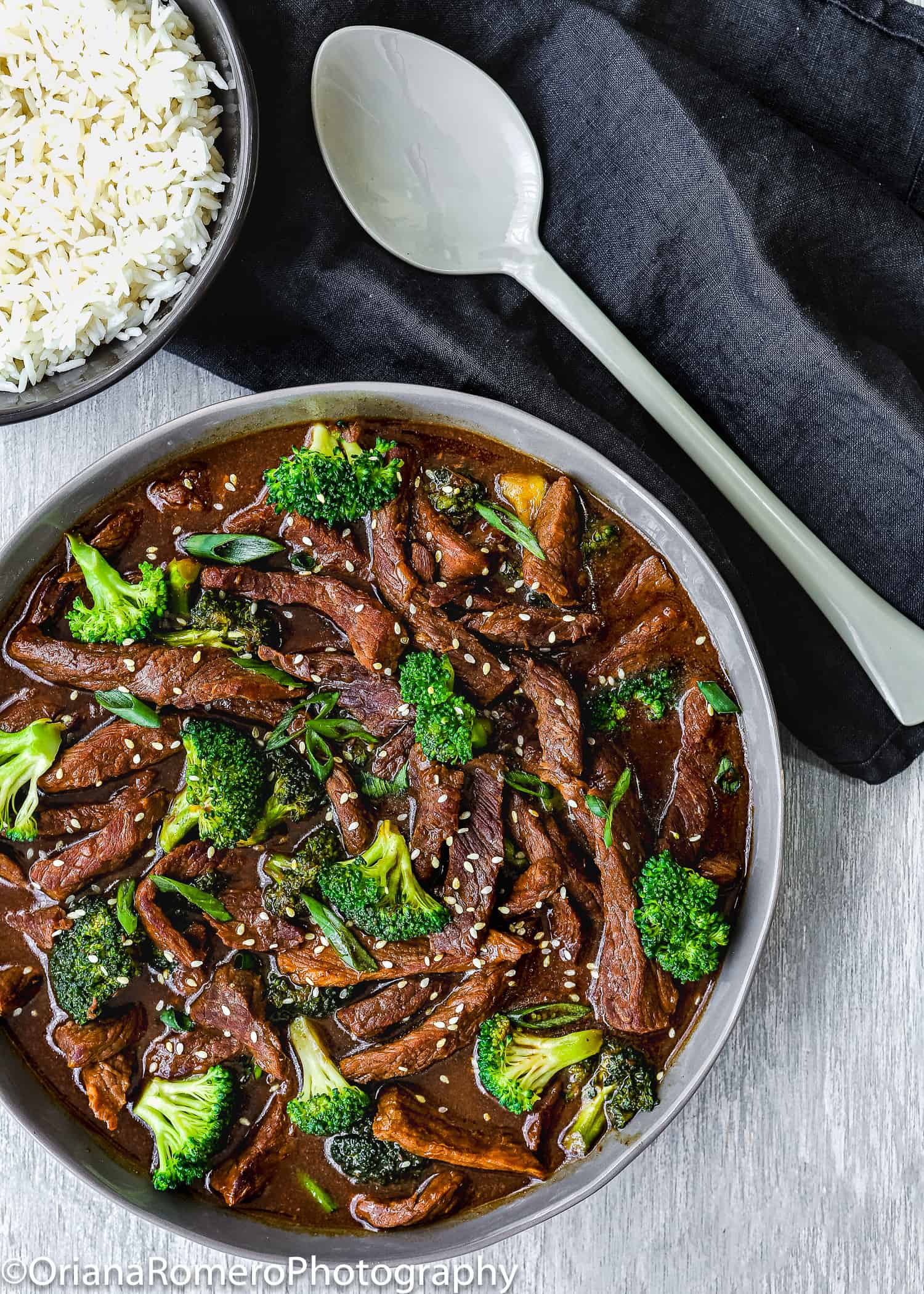 Beef and broccoli stir fry garnished with sesame seed in a serving bowl with white rice and a serving spoon on the side.