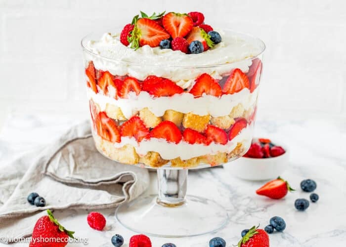A glass trifle dish filled with layers of whipped cream, strawberries, blueberries, and sponge cake, this Eggless Berry Trifle Cake is topped with whole fresh berries for a delightful and colorful treat.