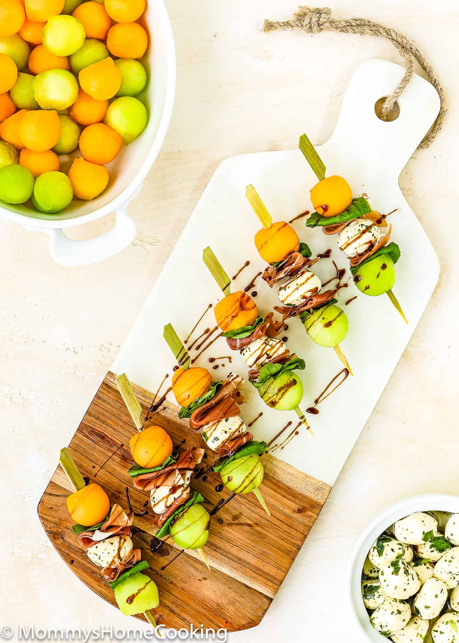 Prosciutto Melon Skewers on a cutting board drizzled with balsamic glaze and melon balls and mini mozzarella on the sides.