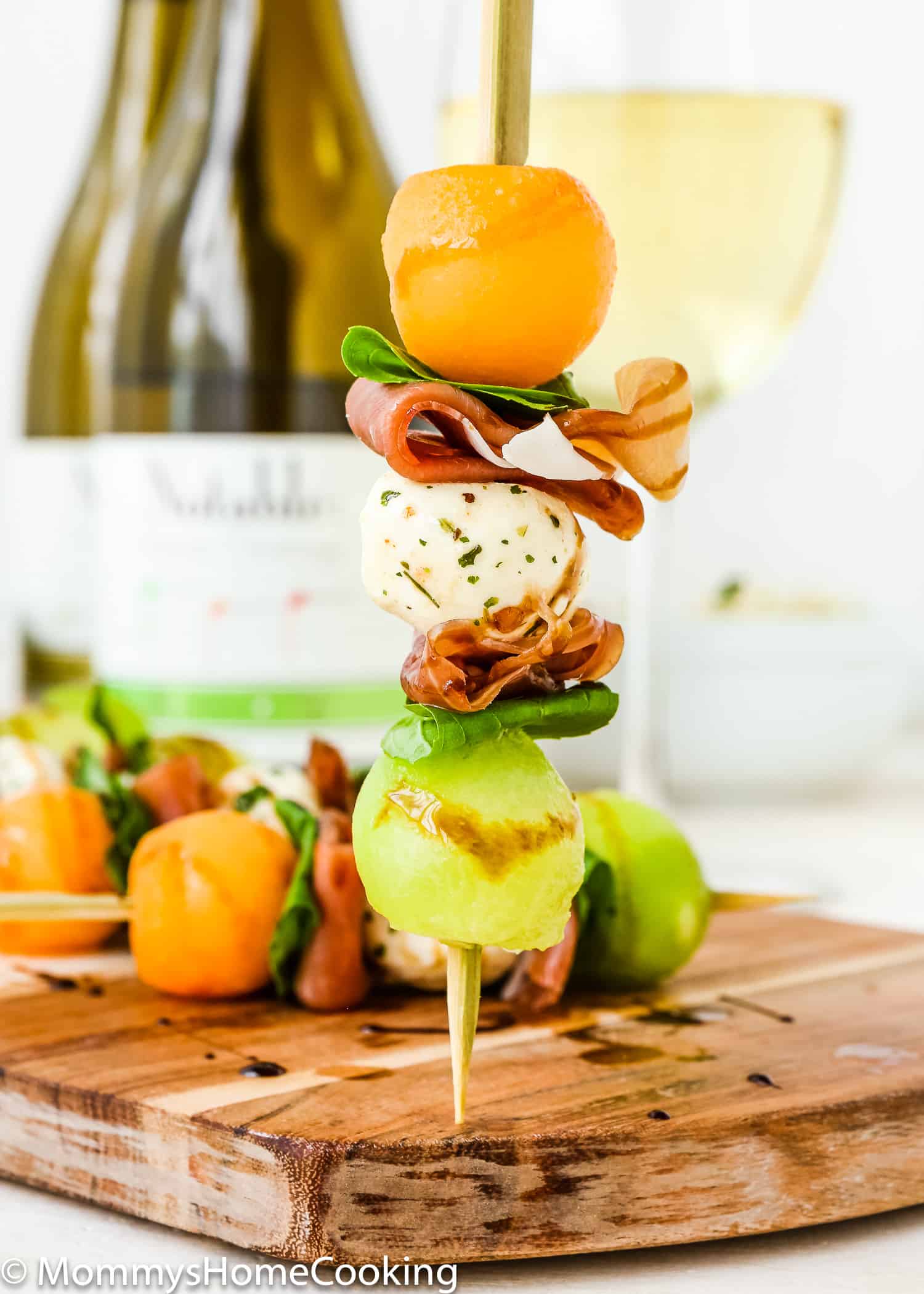 a hand holding a wooden Skewer with cantaloupe, honeydew, Prosciutto and basil leaves.