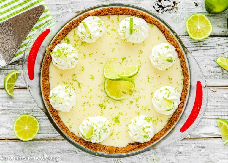 An Easy Eggless Key Lime Pie (No Eggs) with a graham cracker crust, topped with whipped cream, lime slices, and zest sits on a white wooden surface surrounded by fresh limes. This dessert sets perfectly for a creamy, delightful treat.