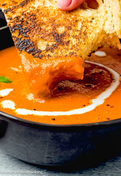 a woman hand holding a grilled cheese sandwich and dip it into a bowl with homemade tomato soup.