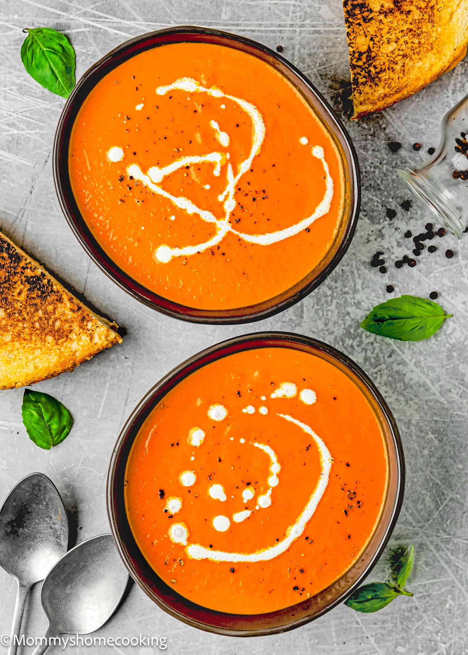 two bowls with Easy Tomato Soup drizzled with cream a black pepper with two grilled sandwiches, basil leaves, and spoons on the side.