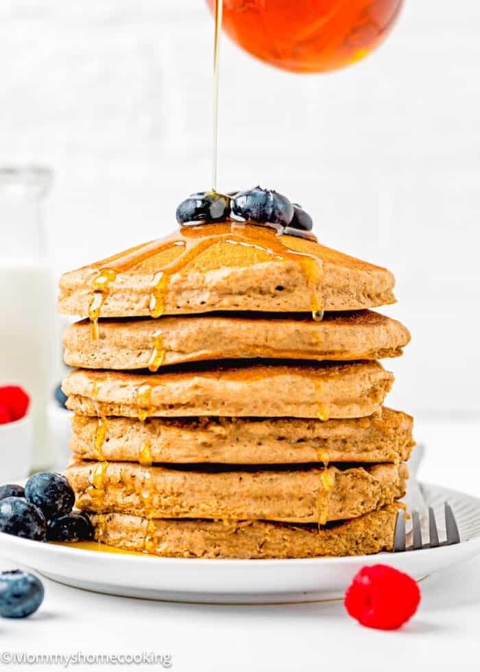 Whole Wheat Pancakes stack with fresh blueberries on top on a plate with maple syrup being poured.