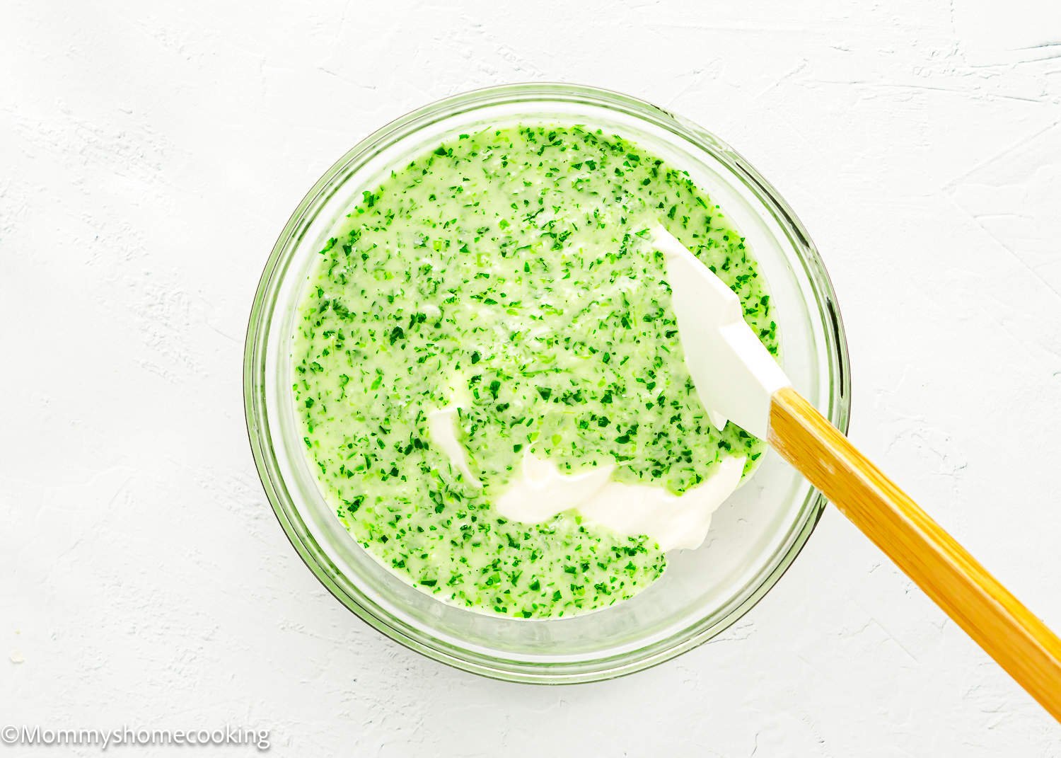 Mayonnaise and veggie mix being mix in a glass bowl with a spatula.