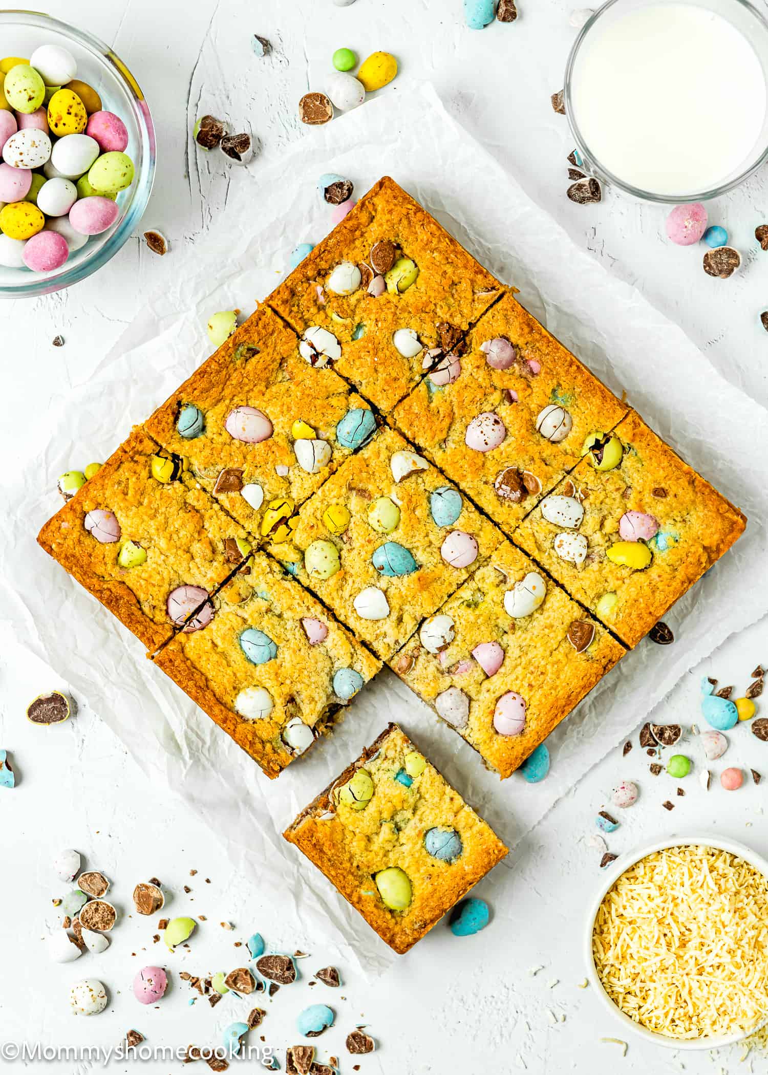 cut into square Easter Blondies over a white surface, with a glass of milk, coconut and easter chocolate eggs.