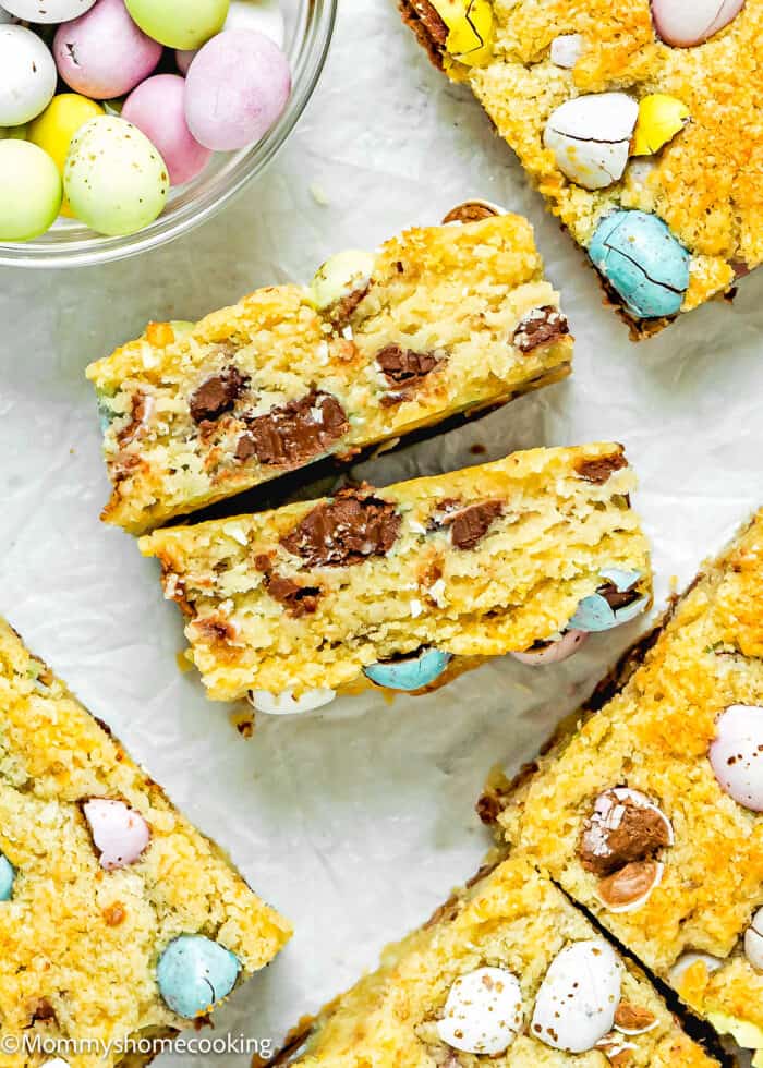 Easter Blondie cut in half showing its inside texture over a white surface with more blondies and chocolate around it.