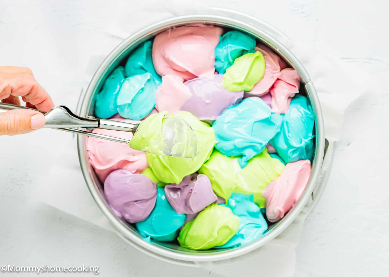 a hand dropping colourful spoonfuls of cheesecake batter in a springform pan.