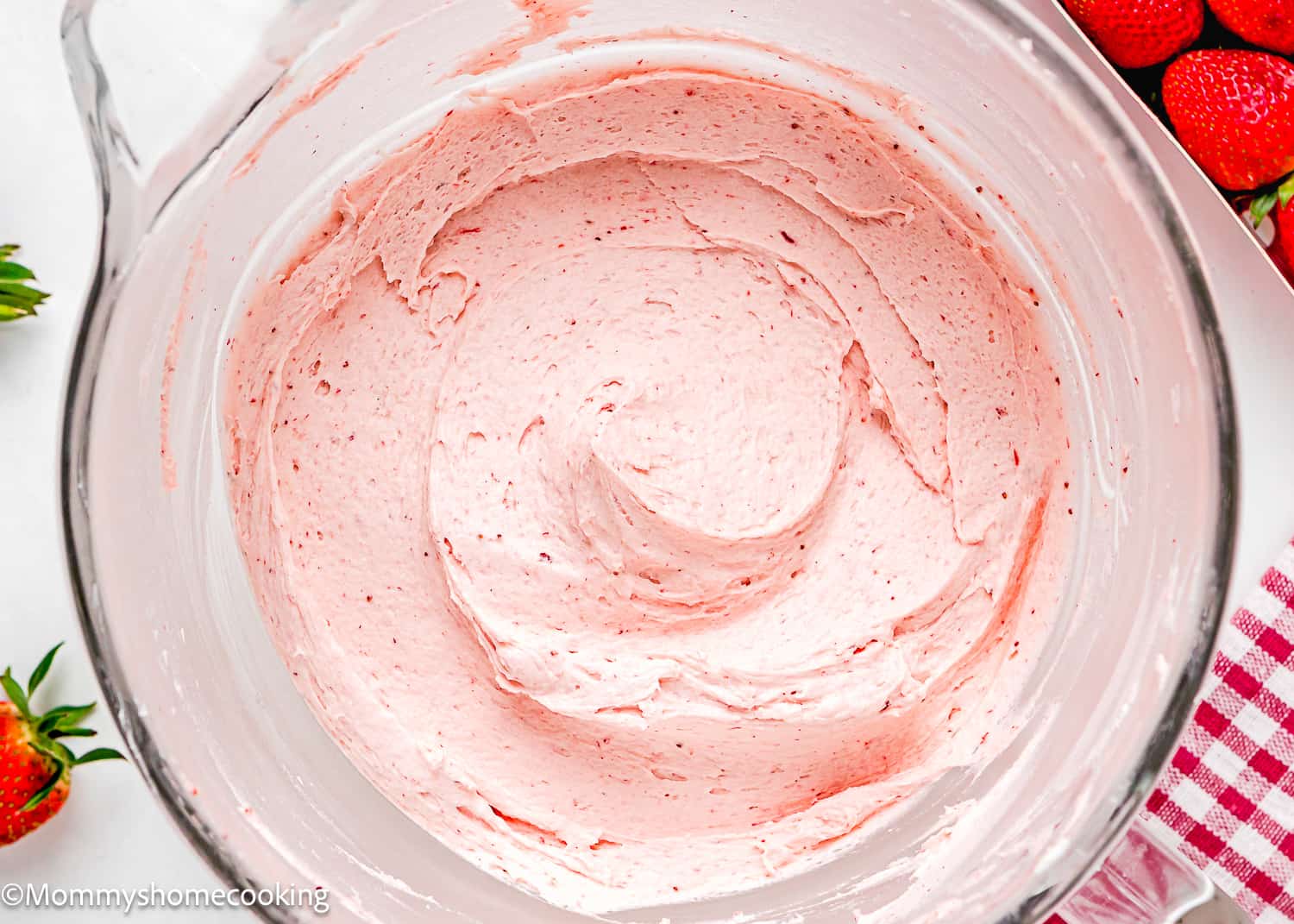 closeup of easy homemade strawberry frosting in a stand mixer bowl over a white surface with fresh strawberries in the side and a red kitchen towel.