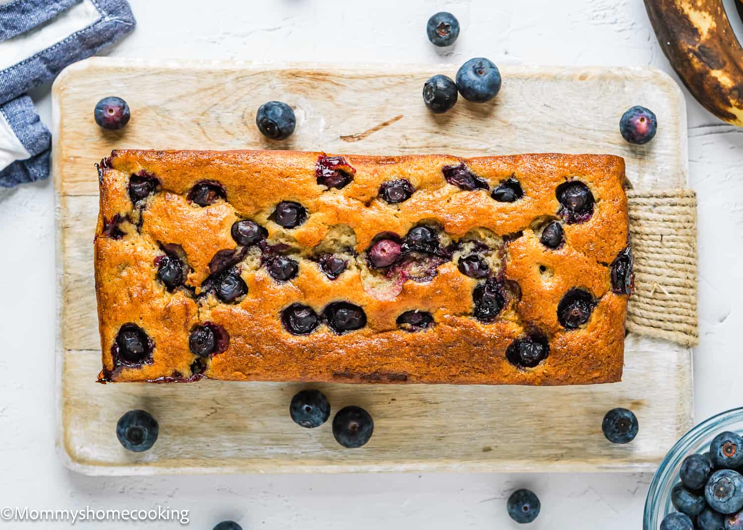 whole Moist Blueberry Banana Bread without eggs over a wooden board with fresh blueberries around it.