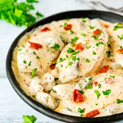 One Pan Creamy Chicken and Mushrooms garnished with chopped Parsley over a blue surface with serving spoons in the background.