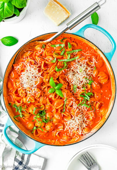 Creamy Tomato Pasta in a blue skillet with a serving spoon, grated cheese basil and a kitchen towel, plates and forks on the side.