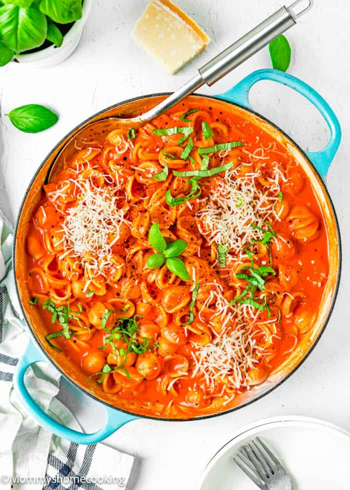 Creamy Tomato Pasta in a blue skillet with a serving spoon, grated cheese basil and a kitchen towel, plates and forks on the side.