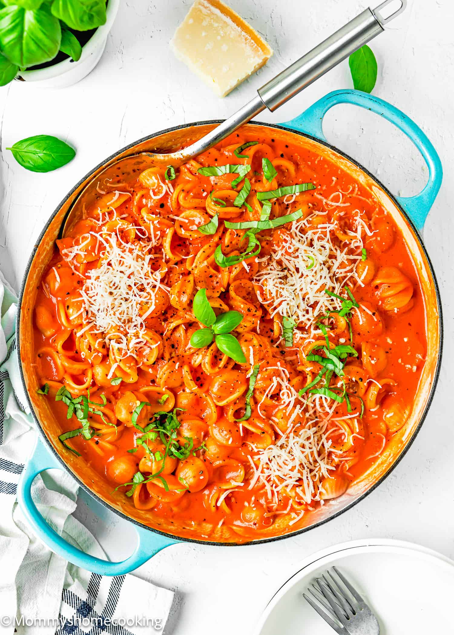 Creamy Tomato Pasta in a blue skillet with a serving spoon, grated cheese basil and a kitchen towel, plates anf forks on the side.
